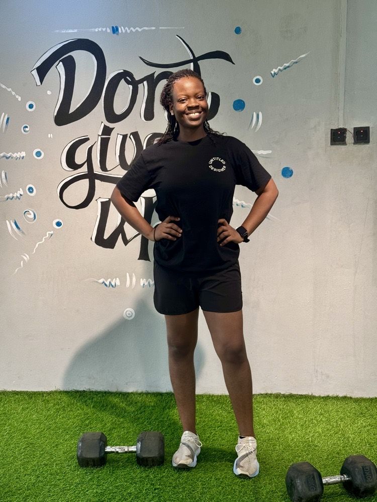 Nana-Fatimah at the gym, hands on hips, in front of a mural that reads Don’t give up